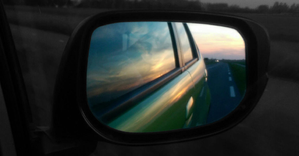 Rückblick im Seitenspiegel eines grünen Autos auf den Sonnenuntergang und die Straße